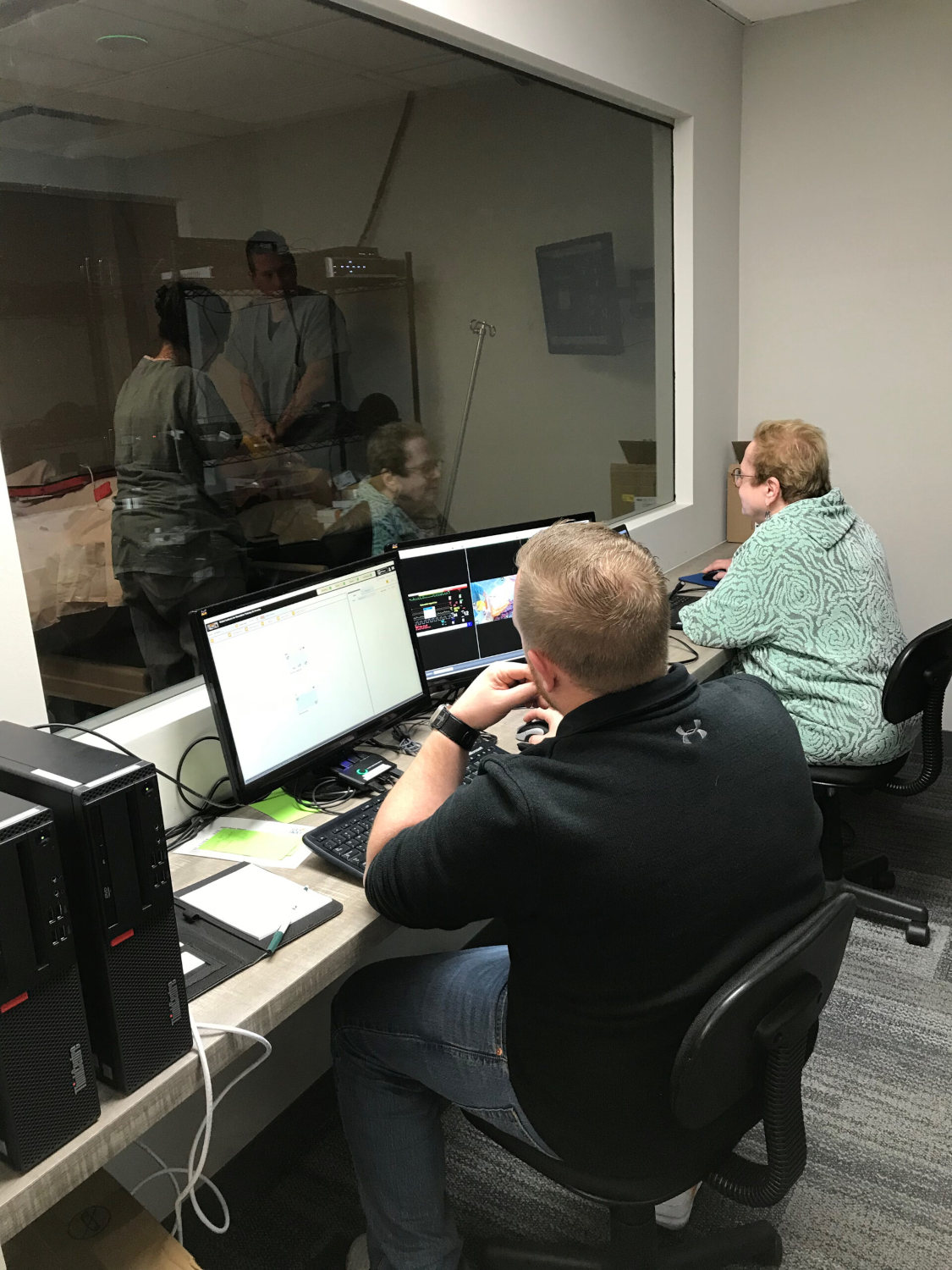 Two technicians sit at a desk with multiple monitors, looking through a glass wall into a room where medical staff are working.