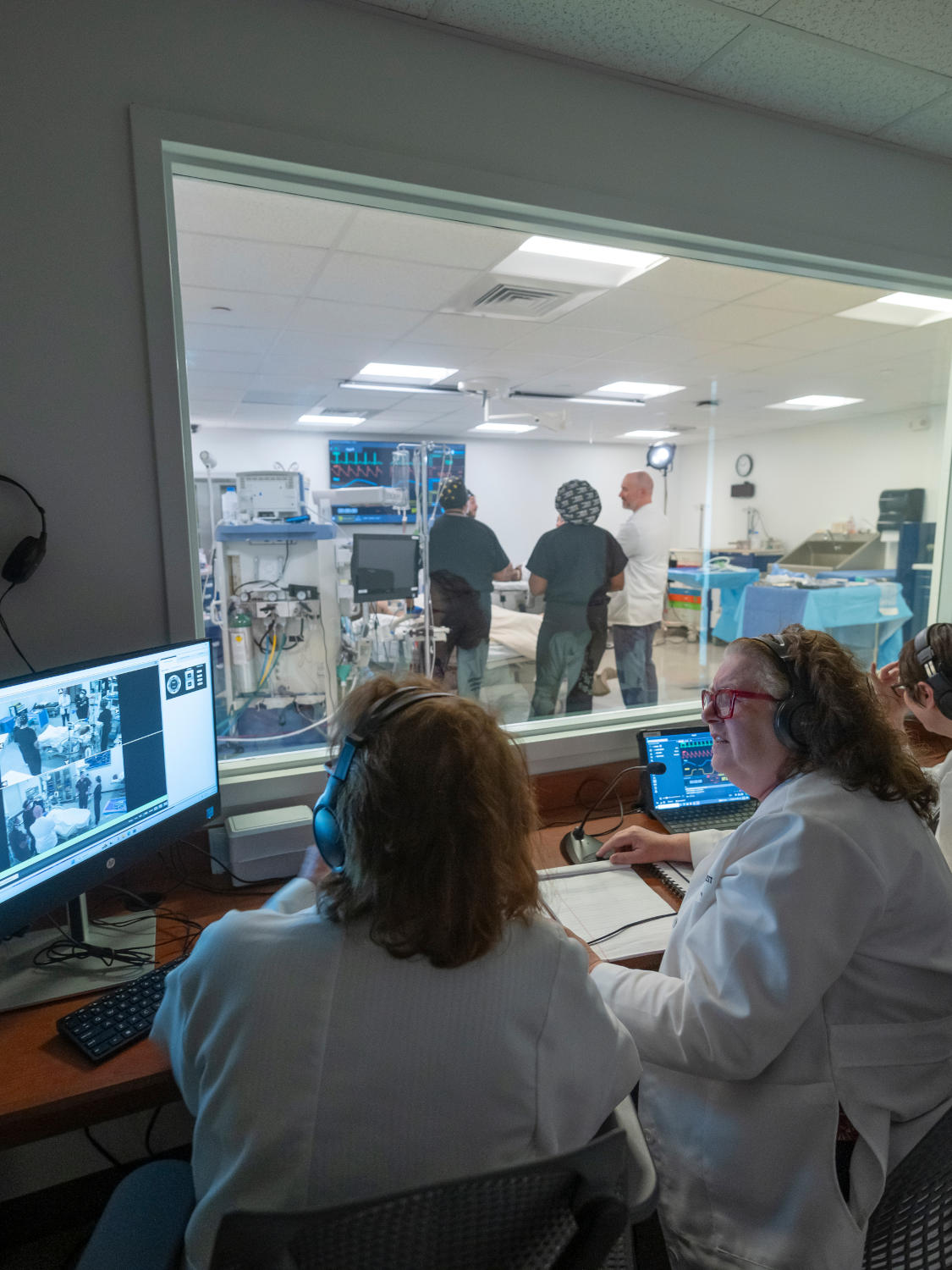 Two clinicians in a control room wearing headsets monitor a live feed as a surgical team works in a bright operating room visible through a window behind them.