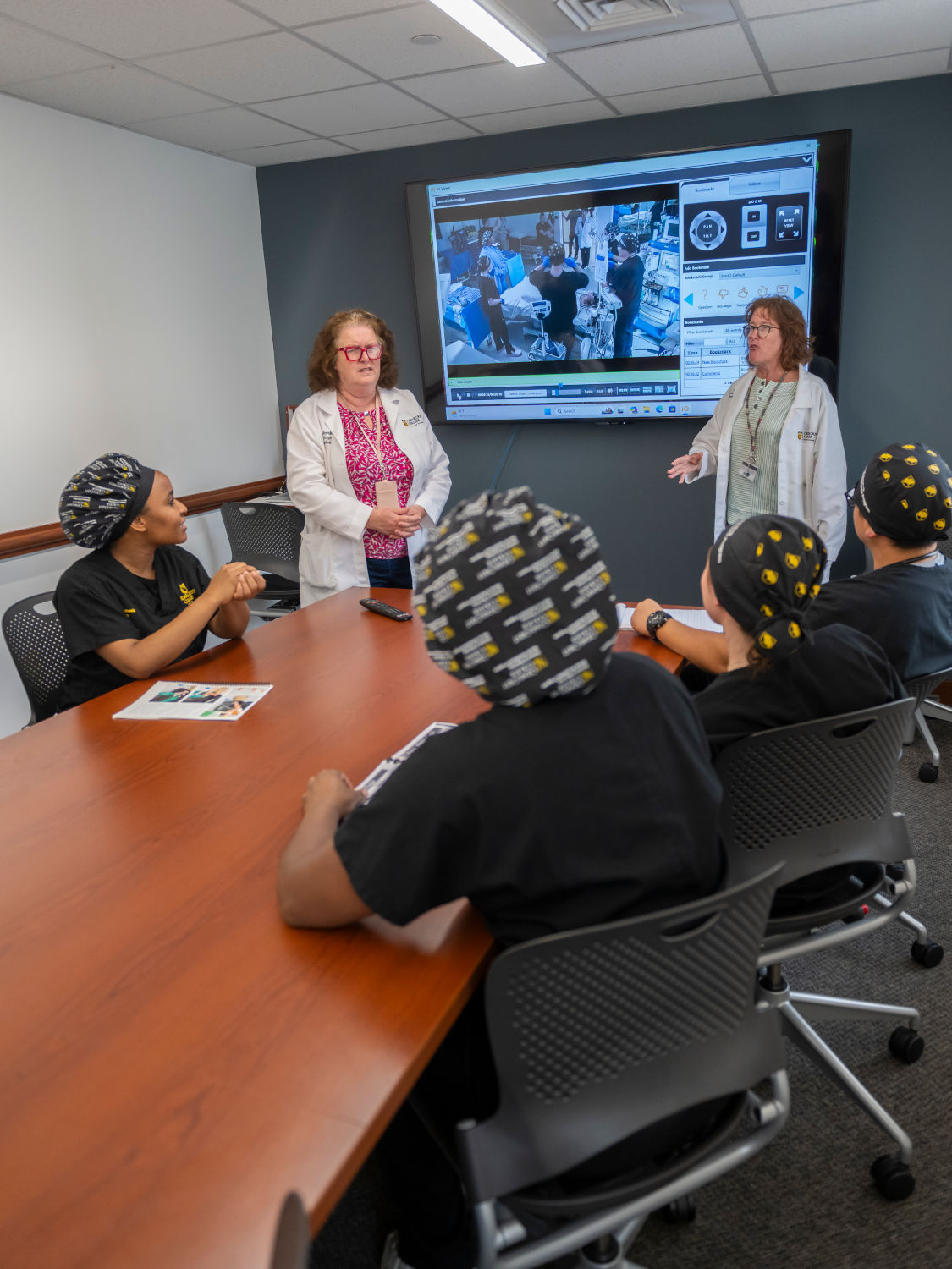 Medical instructors in white coats present a computer display to a classroom of students wearing scrubs and head coverings around a conference table.